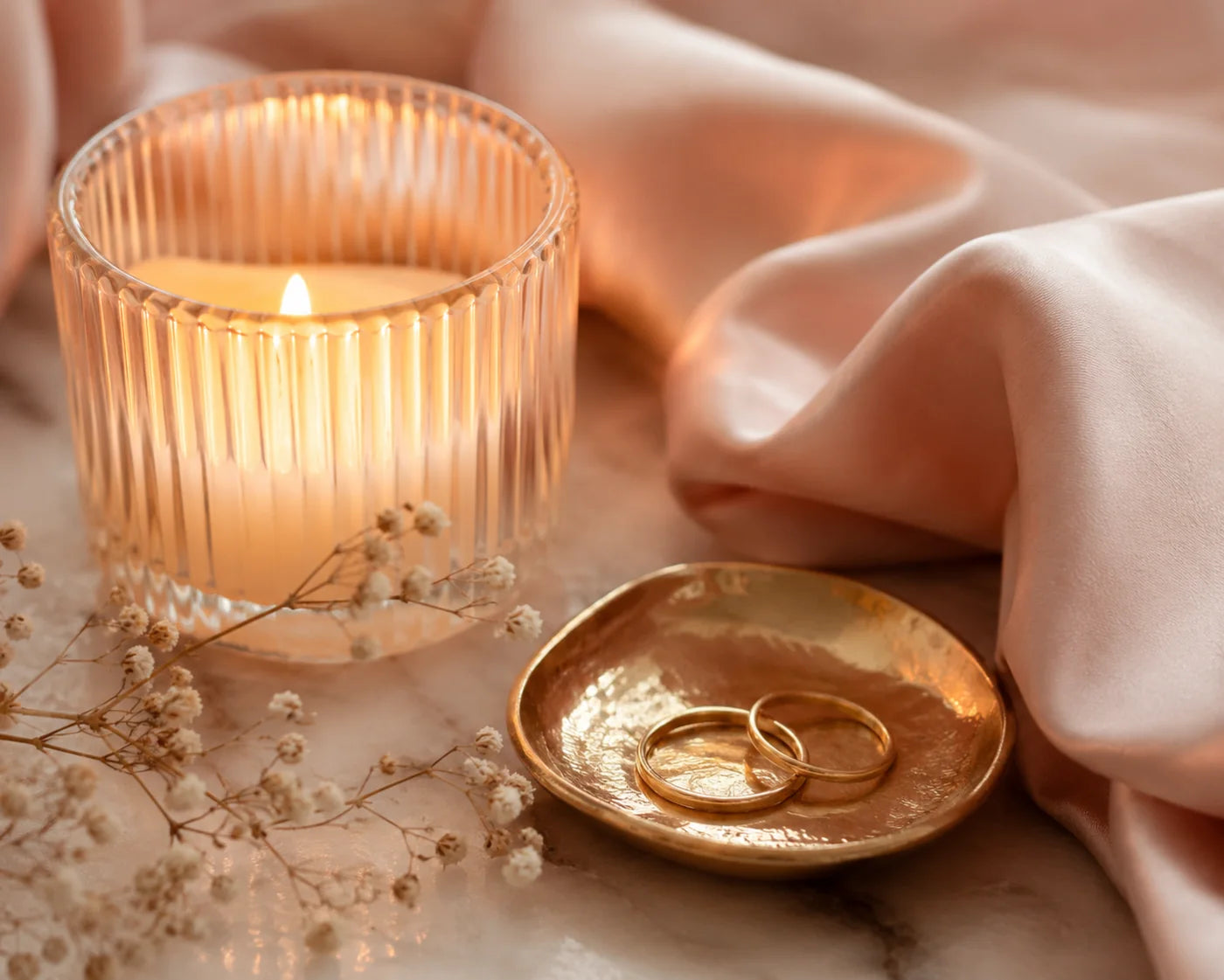 A lit ribbed-glass candle beside blush silk, a gold ring dish with two gold bands and dried baby's-breath